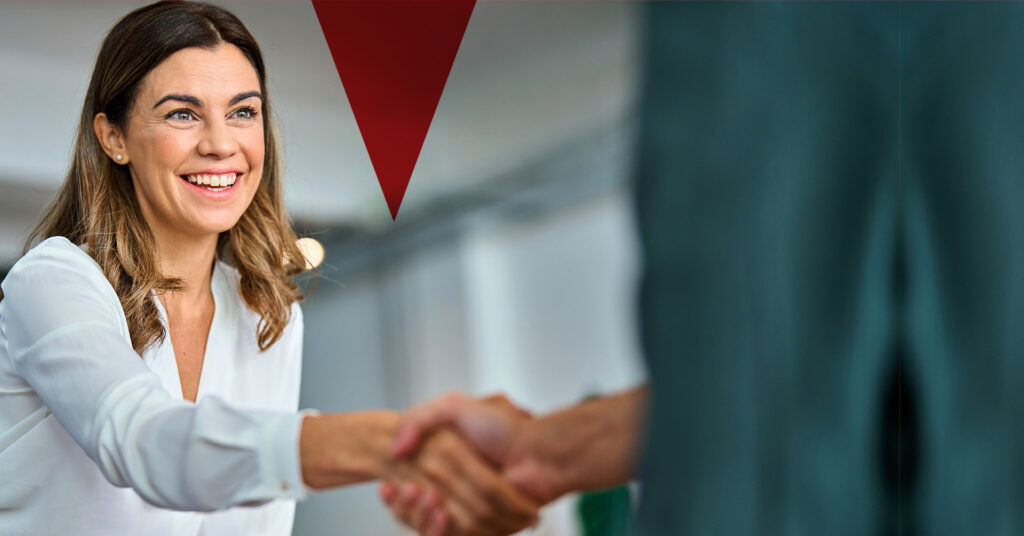 Lächelnde Frau mit braunen Haaren und weißer Bluse gibt einer Person im Vordergrund die Hand, unscharfer Hintergrund mit rotem grafischen Element oben